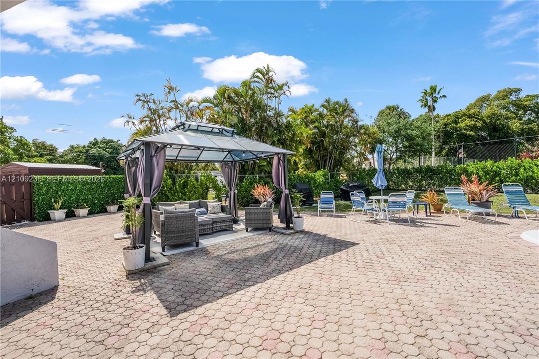 Expansive paver patio featuring a permanent metal-framed gazebo, privacy hedge landscaping, and perimeter fencing