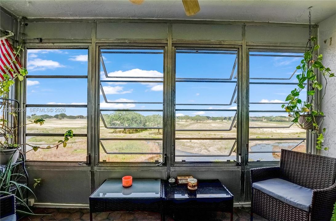 Enclosed patio featuring multi-panel jalousie windows, a textured stucco wall finish, a stone tile floor, and a ceiling-mounted fan