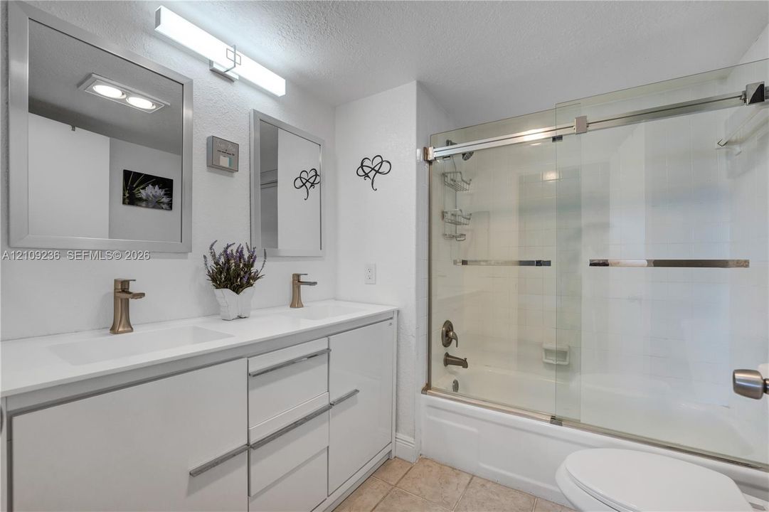 Dual-sink vanity with white cabinetry and solid surface countertop, metallic finish faucets, framed mirrors, and a glass-enclosed shower-tub combination