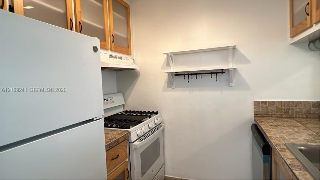 Kitchen featuring wooden cabinetry with glass-panel inserts, tile countertops, white gas range, and built-in wall-mounted shelving