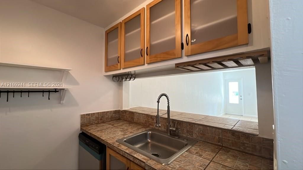 Kitchen area featuring tile countertops, stainless steel sink with high-arc faucet, wood-framed cabinetry with frosted glass inserts, and built-in stemware rack