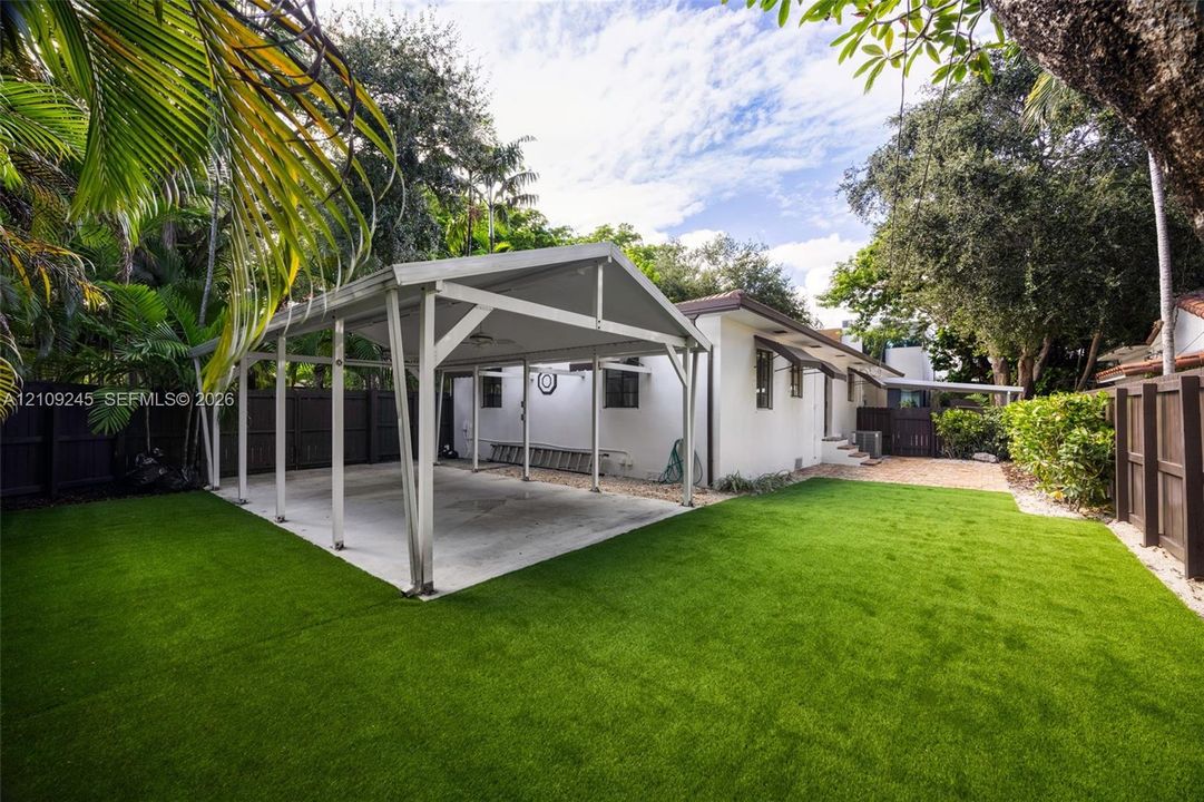 Backyard featuring a concrete-pad carport, synthetic turf lawn, dark wood-finish privacy fencing, and mature palm trees