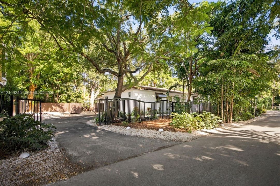 Gated entrance featuring a paved driveway, metal perimeter fencing, mature tree canopy, and landscaped stone borders