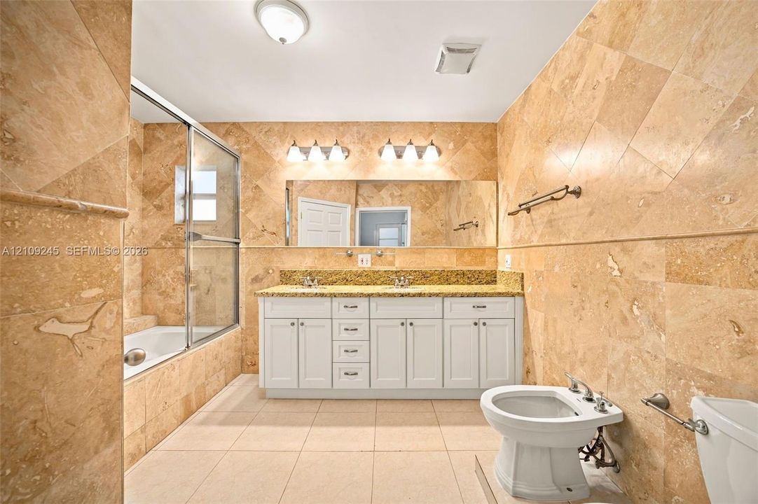 Double vanity with stone countertop, full-width mirror, tile-surround soaking tub with glass enclosure, and floor-to-ceiling wall tile