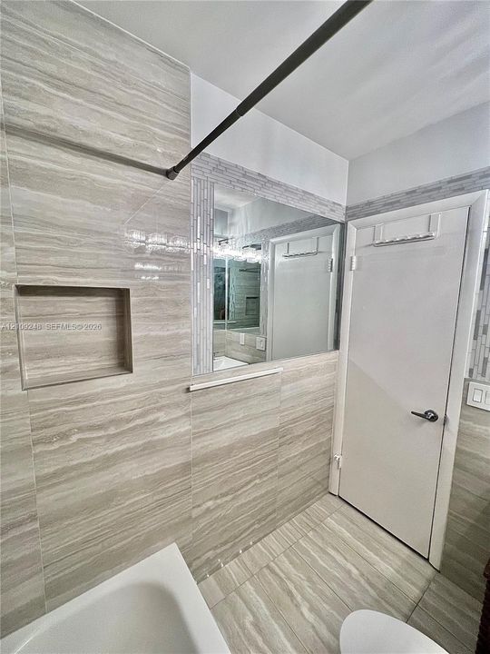 Bathroom featuring neutral-toned stone-finish wall and floor tile, recessed wall niche, glass-fronted medicine cabinet, and decorative linear mosaic tile trim