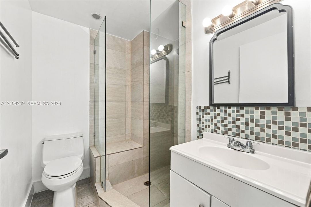 Bathroom featuring a frameless glass shower enclosure with built-in tiled bench, walk-in shower with neutral stone-finish tile, integrated sink vanity with multi-colored mosaic backsplash, and decorative wall-mounted mirror