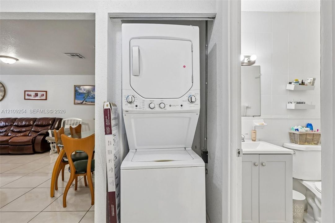 Laundry closet featuring a white stacked washer and dryer unit, cabinet vanity with a rectangular sink, wall-mounted shelving, and tile flooring