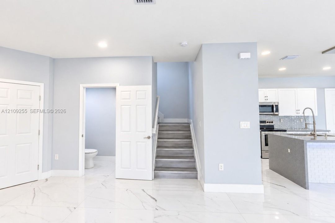 Large-format white tile flooring with grey veining, staircase with wood-finish steps, and a kitchen featuring white cabinetry, a center island with a stone-finish countertop, and stainless steel appliances