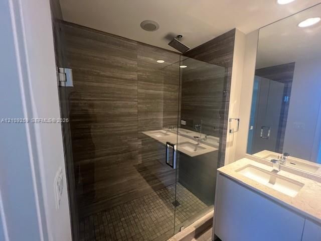 Walk-in glass-enclosed shower featuring horizontal-patterned tile, rain-style showerhead, mosaic tile flooring, and dual-sink vanity with a solid-surface countertop