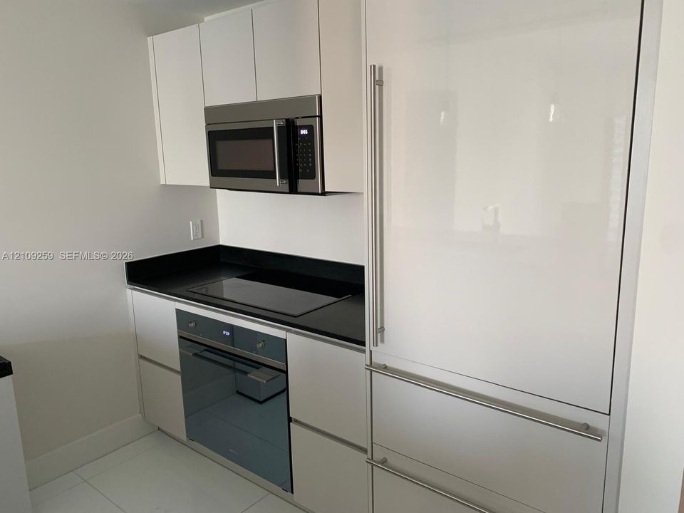 Modern kitchen featuring white flat-panel cabinetry, dark stone countertops, stainless steel built-in microwave, integrated refrigerator, and built-in electric cooktop with wall oven