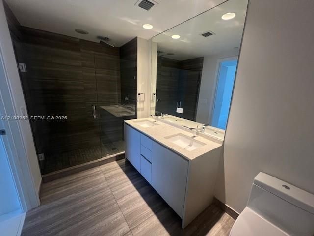 Dual-sink vanity with a solid-surface countertop, walk-in shower with floor-to-ceiling tile, full-width wall mirror, and large-format wood-finish tile flooring