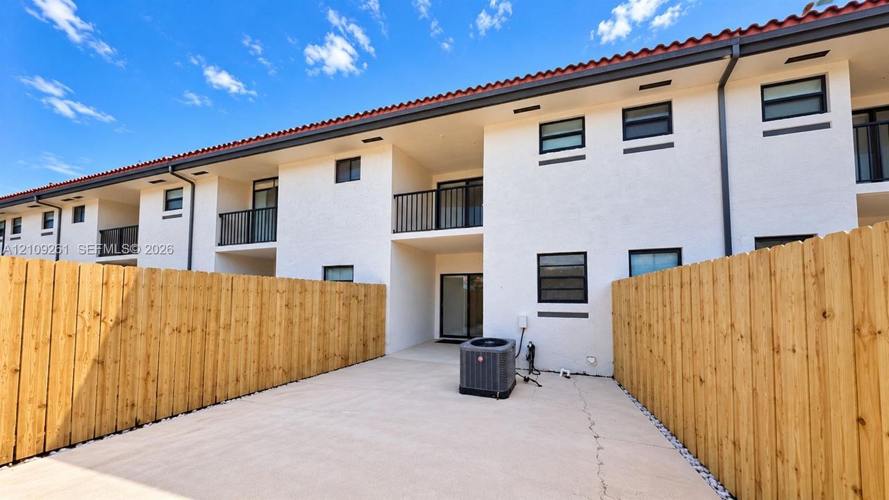 Stucco exterior building with terracotta-tiled roof, second-story metal-railed balconies, private concrete patio, and natural wood privacy fencing
