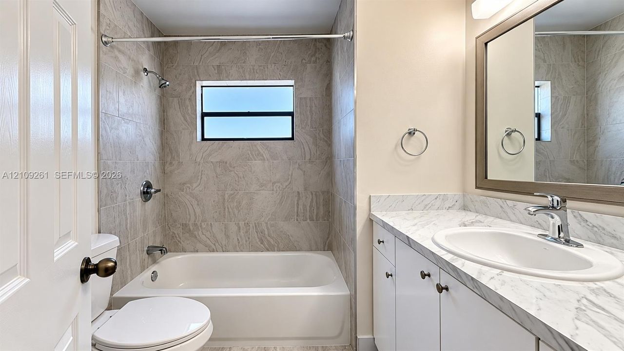 Full-height neutral stone-style tile shower surround, integrated white vanity with stone-patterned countertop, undermount sink, and polished chrome fixtures
