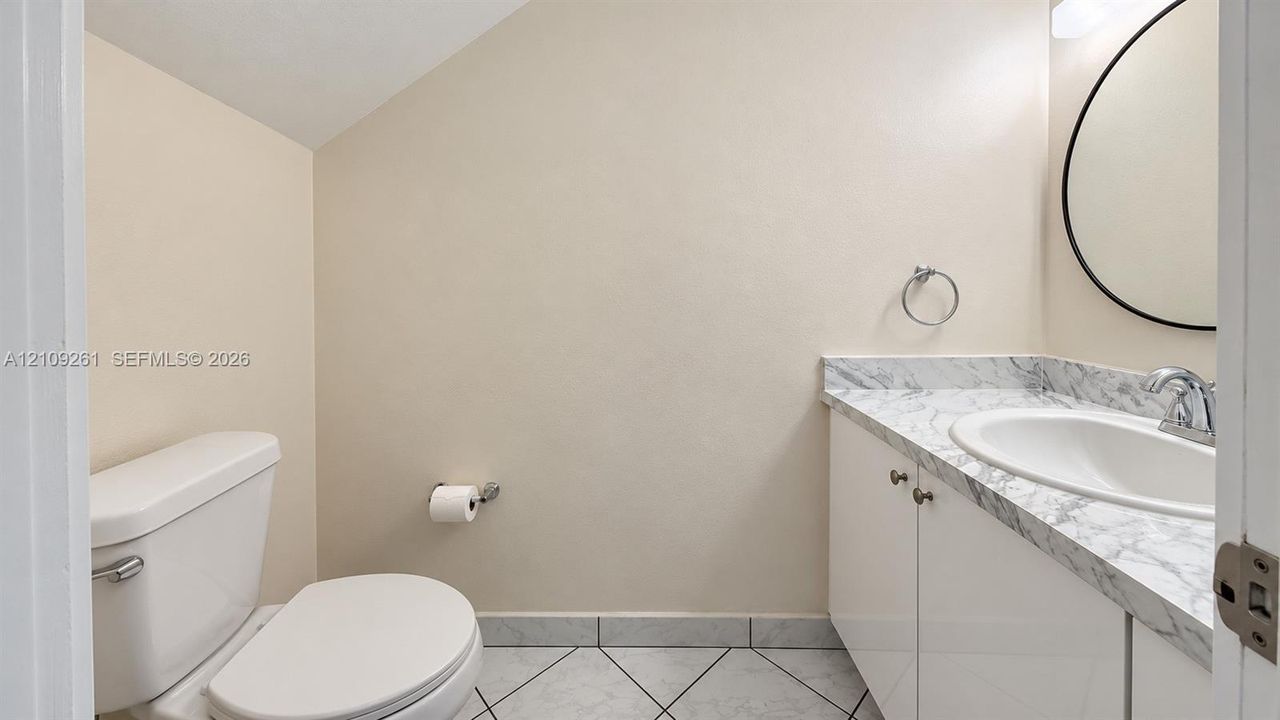 Powder room featuring a vanity with stone-finish countertop and oval basin, circular wall-mounted mirror, white cabinetry, and ceramic tile flooring