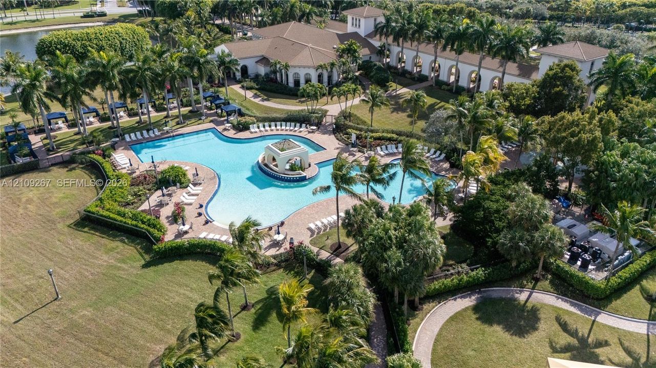 Extensive swimming pool with a central cabana structure, paved pool deck, manicured lawns, mature palm trees, and terracotta-tiled roof architecture