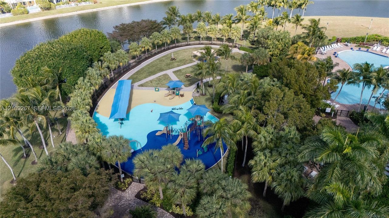 Waterfront community recreation area featuring a multi-tiered swimming pool, shaded splash pad, paved walking paths, and mature palm landscaping