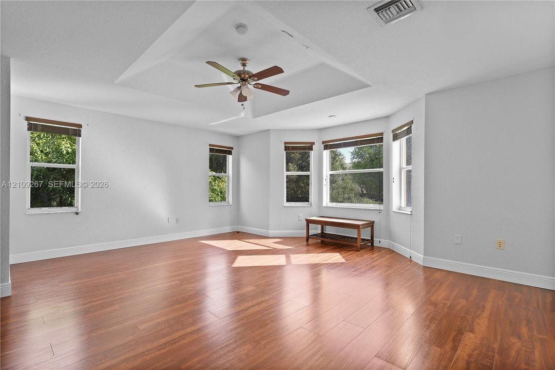 Tray ceiling featuring a central multi-blade ceiling fan, expansive wood-finish flooring, and a multi-window bay area