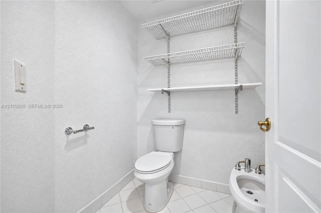 White porcelain toilet, bidet with metal hardware, tiled flooring, and adjustable wall-mounted wire shelving
