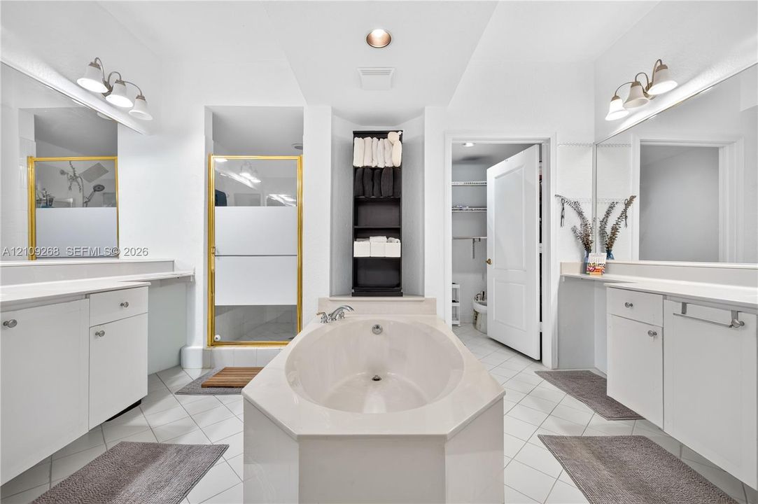 Bathroom featuring dual white vanities with solid-surface countertops, a center soaking tub, a glass-enclosed shower with metallic framing, and tile flooring