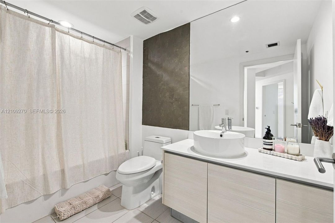 Bathroom featuring a vanity with wood-finish cabinetry, a white countertop, and a vessel sink, accompanied by a full-wall mirror, tiled flooring, and a tub-shower combination with a large-format tiled wall accent