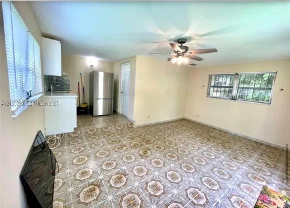 Open-concept area featuring patterned tile flooring, a stainless steel refrigerator, white cabinetry, a ceiling fan with integrated lighting, and horizontal blinds