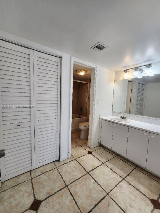 White vanity with an integrated basin, wall-to-wall mirror, louvered-style bi-fold closet doors, and ceramic tile flooring with dark accent inlays
