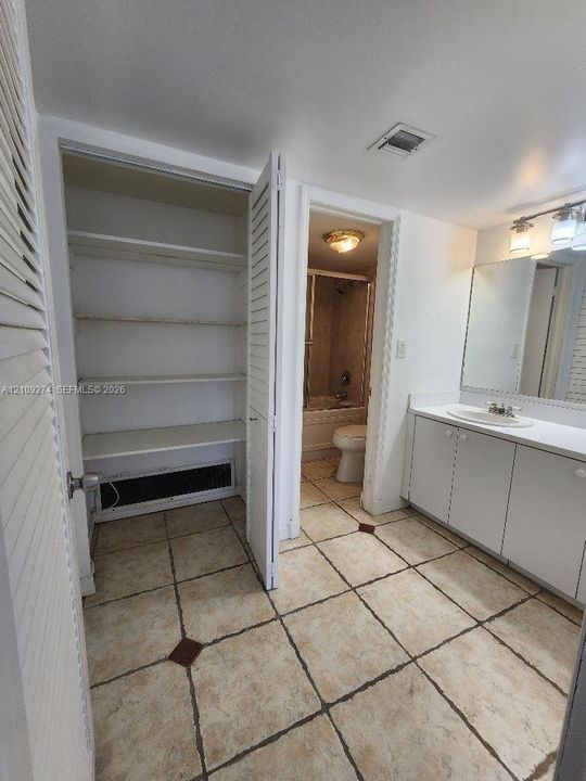 Ceramic tile flooring with decorative inlay accents, vanity with integrated basin, large wall-mounted mirror, three-light fixture, and built-in linen storage with louvered doors