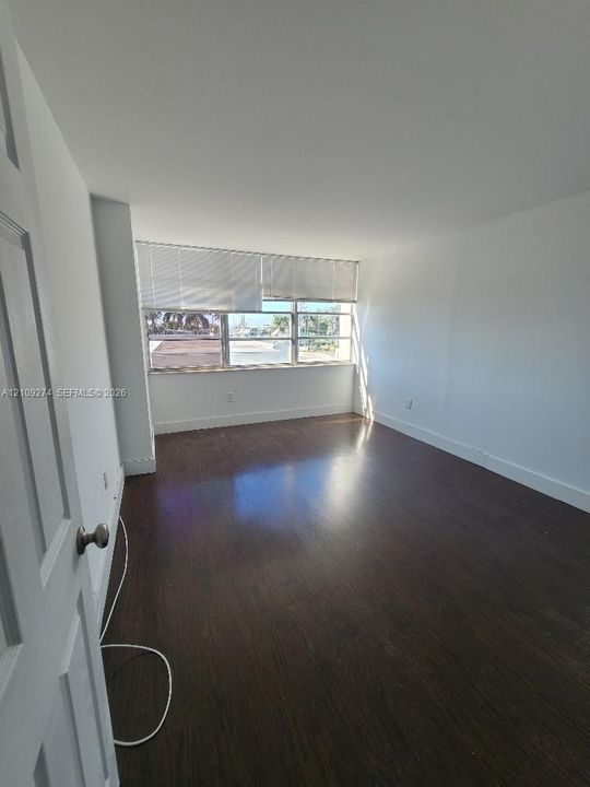 Room featuring dark-toned wood-finish flooring, white walls, and a multi-pane window with adjustable blinds