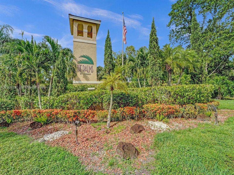 Stucco community monument with architectural tower detail, surrounded by manicured tropical landscaping, mature palm trees, dense hedging, and decorative stone mulch beds