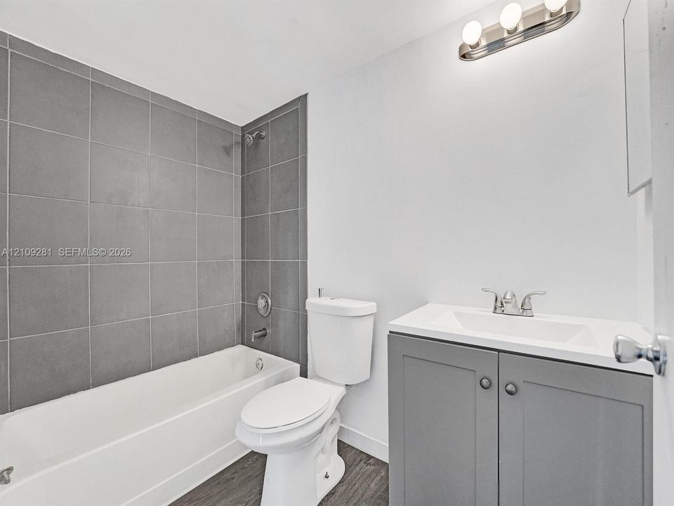 Bathroom featuring a grey-tiled shower surround, vanity with a white integrated basin, brushed nickel faucet, wood-finish flooring, and multi-bulb wall lighting