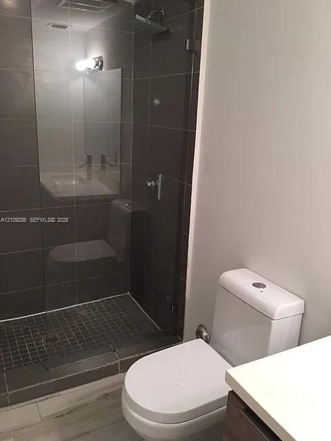 Walk-in shower with dark wall tiling, glass enclosure, and rainfall showerhead, accompanied by a dual-flush toilet and light-toned wood-finish flooring