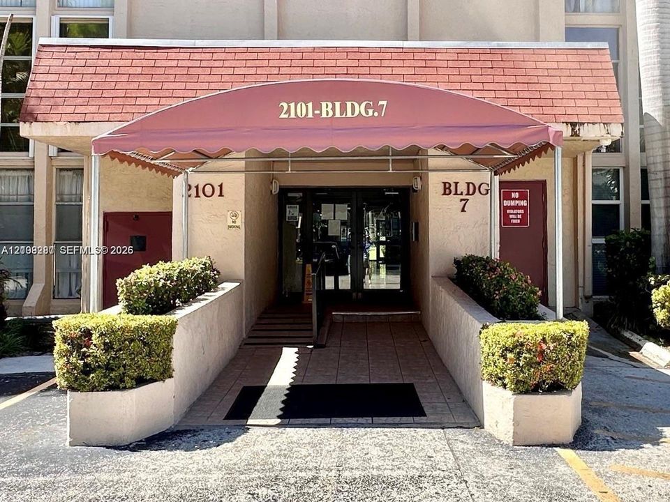 Building entrance featuring a red canvas awning, shingle-roof overhang, paved walkway, and built-in masonry planters