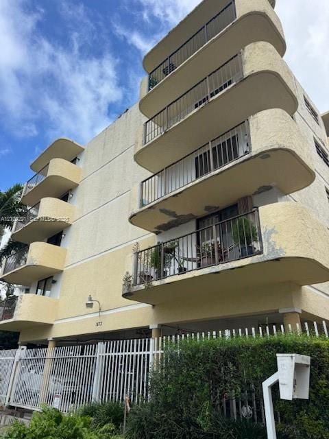 Multi-story stucco exterior featuring rounded, cantilevered balconies with metal railings, a secure gated parking perimeter, and exterior wall-mounted lighting