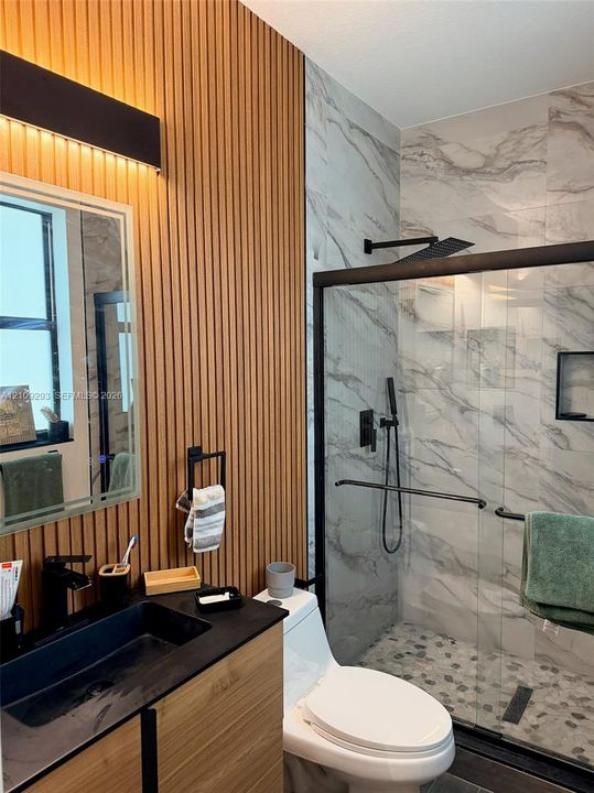 Bathroom featuring a wood-slat accent wall, a floating vanity with a rectangular vessel sink, and a walk-in shower with stone-tile flooring, floor-to-ceiling wall tile, and matte black fixtures