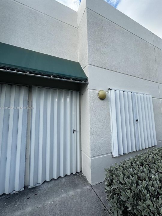 Textured stucco exterior featuring a dark green fabric awning, vertical accordion-style security shutters, and a wall-mounted globe light fixture