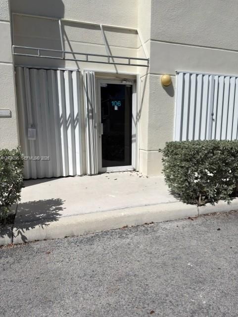 Exterior entryway featuring a glass-panel door, metal accordion hurricane shutters, a structural metal awning, and wall-mounted exterior lighting