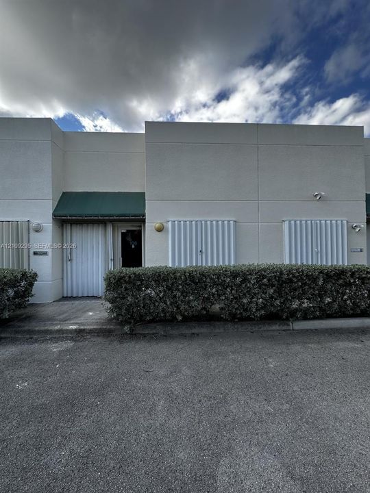 Stucco exterior featuring vertical metal security shutters, a green fabric awning, exterior wall-mounted lighting, and manicured shrubbery