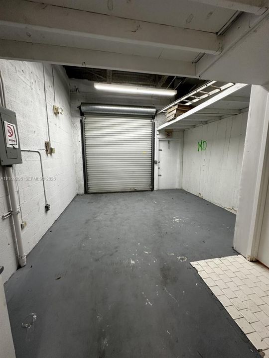 Enclosed garage space featuring a roll-up security door, painted concrete flooring, an overhead fluorescent light fixture, and integrated wall shelving
