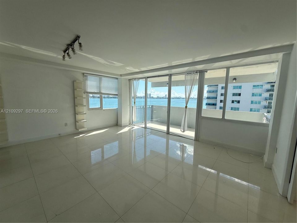 Spacious room featuring light-colored polished tile flooring, floor-to-ceiling glass sliding doors, a built-in wall-mounted shelving unit, and an attached balcony