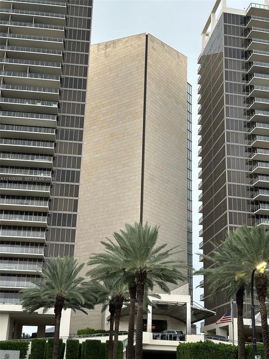 High-rise architectural exterior featuring stone cladding, multi-story glass curtain walls, and stacked private balconies, complemented by mature palm landscaping