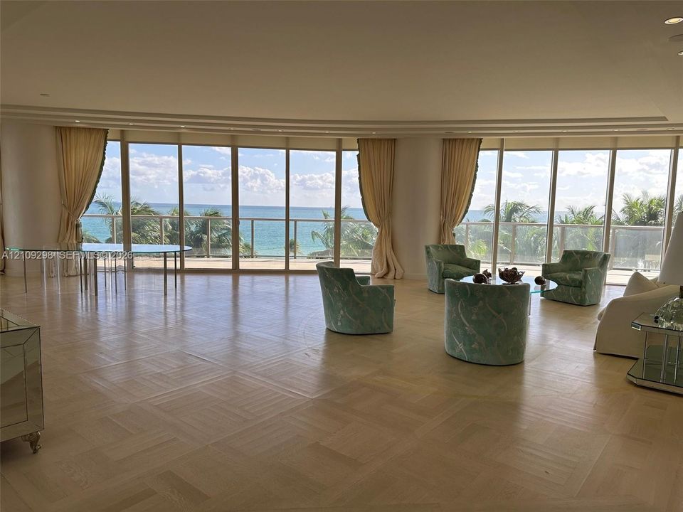 Expansive interior featuring parquet wood flooring, floor-to-ceiling curved glass wall panels, recessed lighting, and a metal-railed balcony with ocean scenery