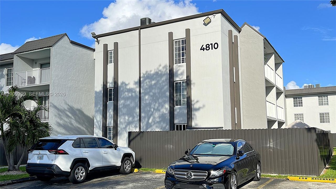 Three-story multi-family structure featuring a smooth stucco exterior, vertical architectural accents, grid-pattern windows, and a metal perimeter fence