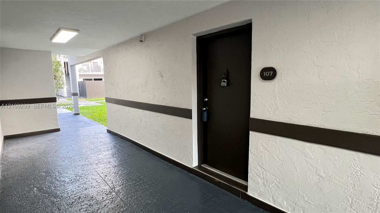 Covered exterior entryway featuring a dark-toned door with a circular unit number plaque, textured light-colored walls with horizontal trim, and painted concrete flooring