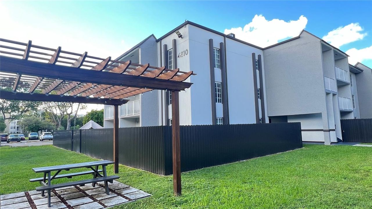 Exterior featuring a multi-story structure with a contemporary facade, wood-frame pergola, metal perimeter fencing, and landscaped grounds