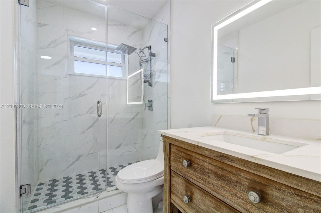 Bathroom featuring a wood-finish vanity with a stone countertop, an integrated rectangular sink, a frameless glass-enclosed shower with large-format tile, and an illuminated wall mirror