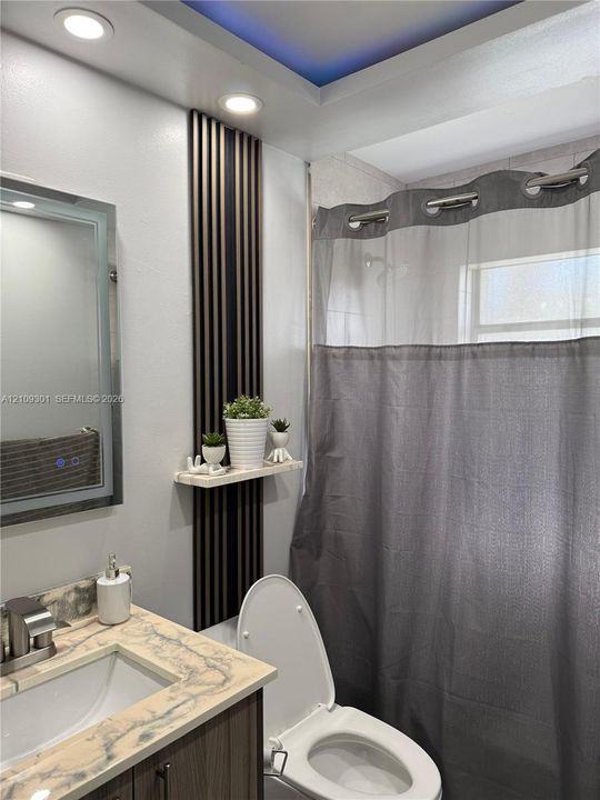 Bathroom featuring a stone-look countertop with an integrated sink, vertical slat accent wall paneling with a floating shelf, recessed ceiling lighting, and a tray ceiling detail with integrated color-accent lighting