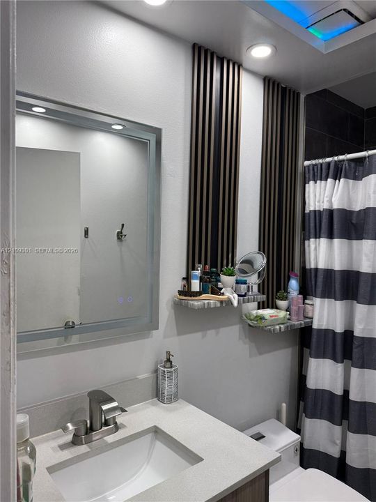 Bathroom featuring a vanity with a stone-surface countertop, an integrated rectangular sink, a polished metal faucet, a backlit rectangular mirror, and decorative wall-mounted acoustic-style slats with built-in stone-finish shelving