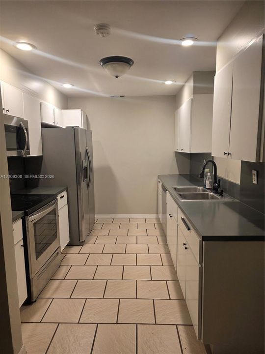 Kitchen featuring white shaker-style cabinetry, stainless steel appliances, dark solid-surface countertops, dual-basin stainless steel sink with a matte black faucet, and tiled flooring