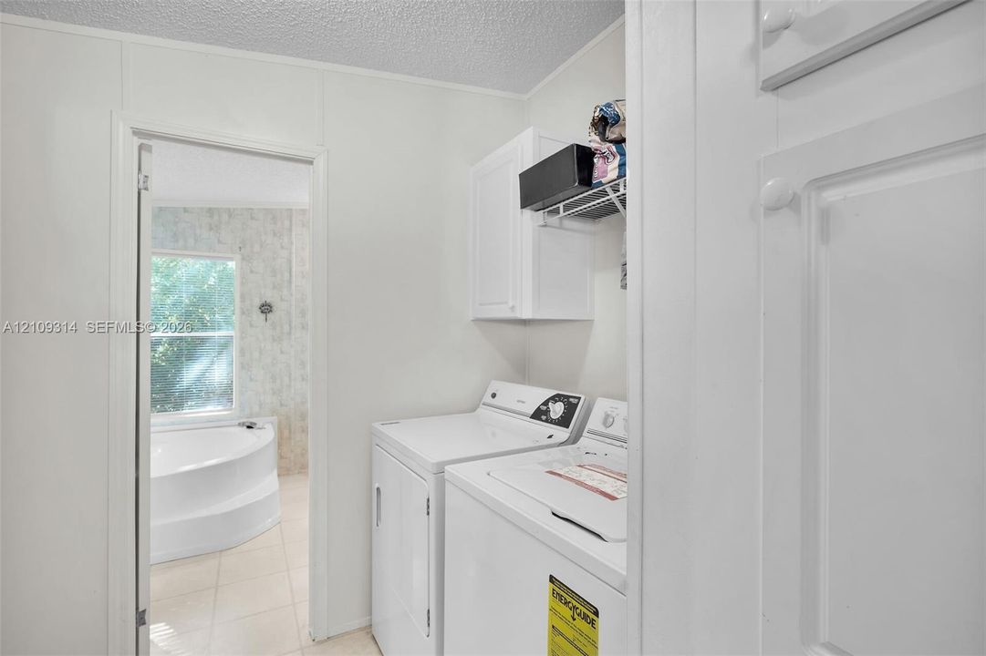 Laundry area featuring white built-in cabinetry, wire shelving, and space for side-by-side appliances, with tile flooring and a soaking tub visible beyond the doorway