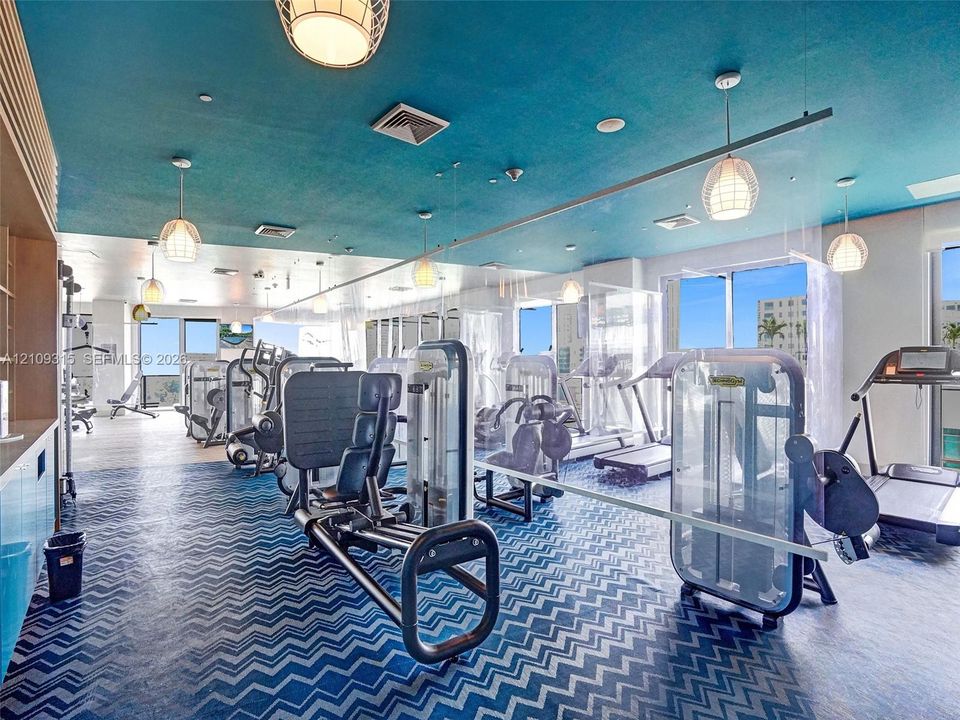 Fitness center featuring herringbone-patterned flooring, bold teal ceiling, large windows, and multiple built-in pendant lights